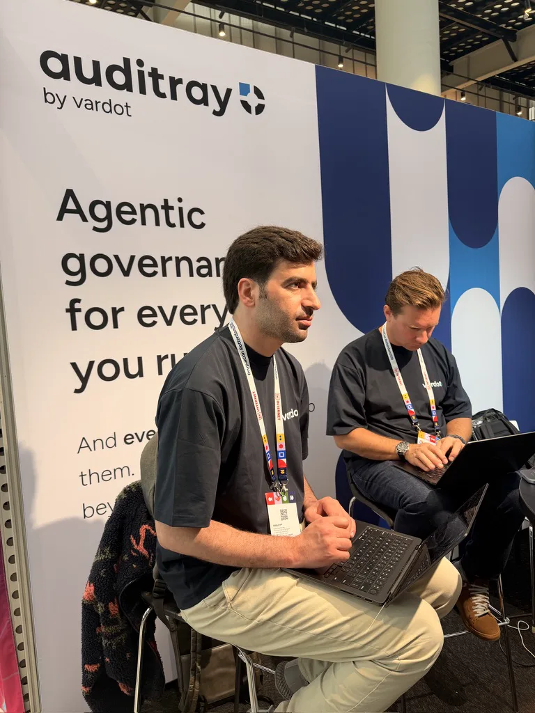 Two Vardot team members working on laptops in front of the Auditray booth wall.