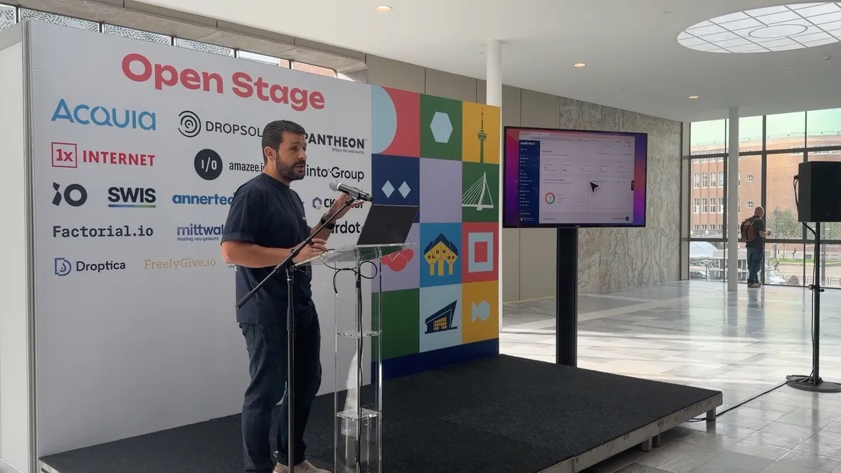 A speaker at a lectern on the DrupalCon Open Stage, beside a screen showing the Auditray dashboard.